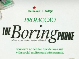 Uma foto de um telefone celular transparente (The Boring Phone) com botões grandes e tela pequena. O telefone está sobre uma mesa de madeira ao lado de uma garrafa de cerveja Heineken. O logo da Heineken e da Bodega estão presentes na imagem.