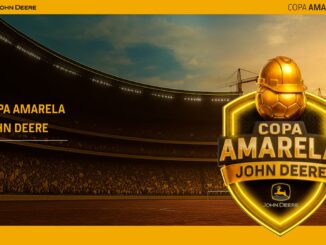 Banner promocional da Copa Amarela John Deere exibindo um grande escudo dourado com uma bola de futebol usando capacete de obra, posicionado à frente de um estádio lotado ao pôr do sol com guindastes de construção ao fundo.