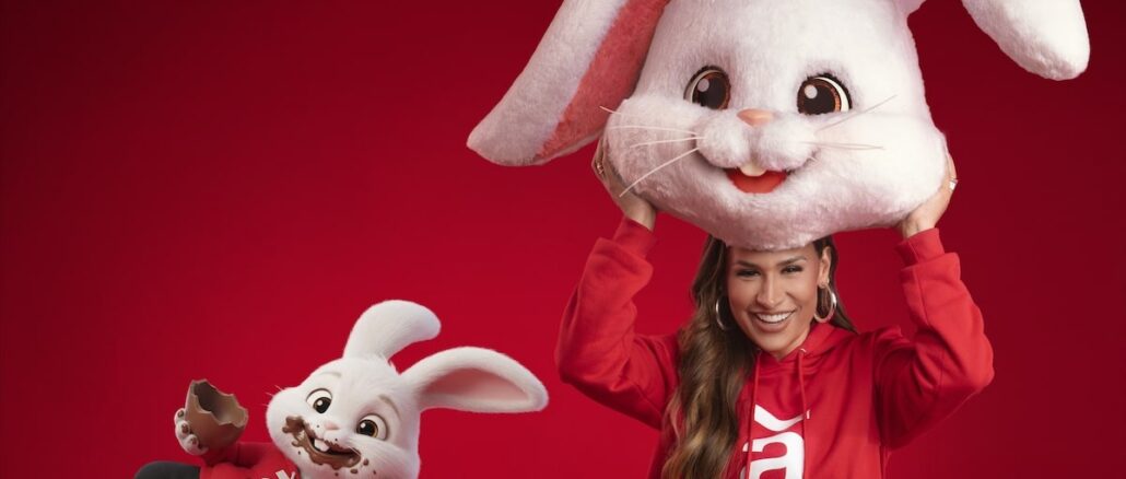 Promoção Páscoa Ameriquinho Americanas 2026 com Simone Mendes e Coelhos de Pelúcia Uma mulher sorridente com moletom vermelho segura uma grande cabeça de mascote de coelho branco sobre si, enquanto um pequeno coelho animado ao lado aparece com o rosto sujo de chocolate e segura um ovo de Páscoa, ambos sobre um fundo vermelho.