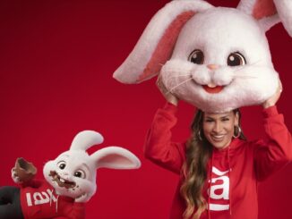 Uma mulher sorridente com moletom vermelho segura uma grande cabeça de mascote de coelho branco sobre si, enquanto um pequeno coelho animado ao lado aparece com o rosto sujo de chocolate e segura um ovo de Páscoa, ambos sobre um fundo vermelho.
