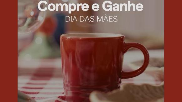 Caneca de ceramica vermelha em destaque sobre uma mesa com toalha xadrez acompanhada pelo texto informativo Compre e Ganhe Dia das Maes.