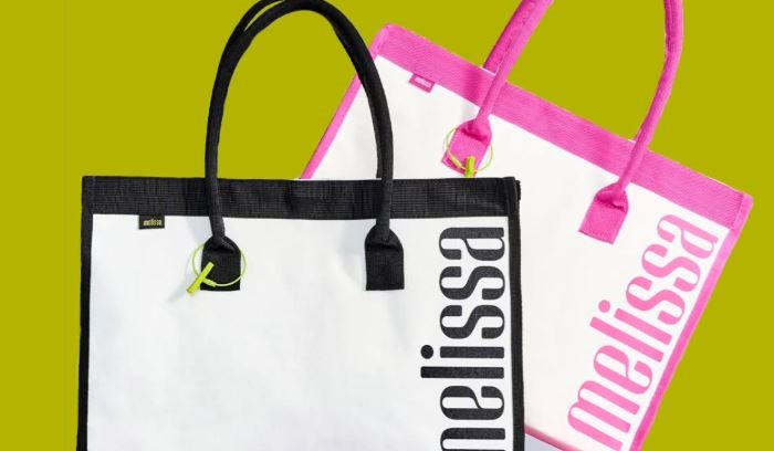 Dia das Mães Melissa Shopper Bag Duas bolsas Melissa Shopper, uma branca com detalhes pretos e outra branca com detalhes rosas, posicionadas sobre um fundo verde.