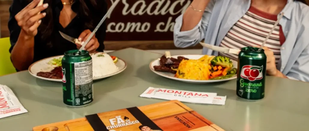 Duas mulheres almoçando em uma mesa de restaurante com pratos de comida, latas de Guaraná Antarctica Zero e embalagens do Montana Grill. Em primeiro plano há um cardápio em formato de tábua de carne.