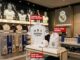 Produtos oficiais do Real Madrid, incluindo camisa branca, boné e mochila, expostos em uma loja moderna com fotos de jogadores ao fundo.