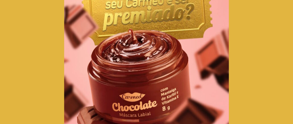 Imagem promocional da máscara labial Carmed Chocolate. O produto está em um pote marrom aberto sobre barras de chocolate, com pedaços de chocolate flutuando ao fundo. No topo, um bilhete dourado traz uma mensagem sobre premiação.