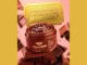 Imagem promocional da máscara labial Carmed Chocolate. O produto está em um pote marrom aberto sobre barras de chocolate, com pedaços de chocolate flutuando ao fundo. No topo, um bilhete dourado traz uma mensagem sobre premiação.