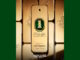 Frasco de perfume dourado em formato de barra de ouro com uma pedra verde central e escrita em árabe cercado por diversos lingotes de ouro sobre um fundo verde.