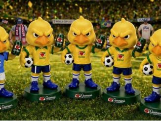 Cinco bonecos colecionáveis do mascote Canarinho Pistola posicionados sobre gramado de estádio segurando latas de Guaraná Antarctica e bolas de futebol.