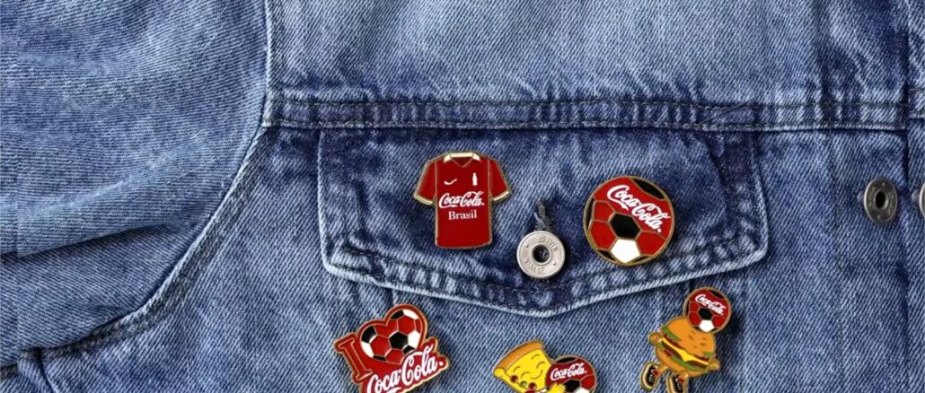 Campanha Coca-Cola Pins Colecionáveis 2026 Cinco pins colecionáveis de esmalte da Coca-Cola com temática futebolística dispostos sobre uma jaqueta jeans, incluindo modelos com camisa do Brasil, bola de futebol, mascote hambúrguer e personagem animado.