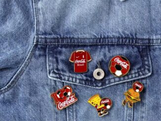 Cinco pins colecionáveis de esmalte da Coca-Cola com temática futebolística dispostos sobre uma jaqueta jeans, incluindo modelos com camisa do Brasil, bola de futebol, mascote hambúrguer e personagem animado.