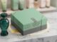 Caixa de presente verde-sage com faixa cinza e texto "Promoção Caixa Premiada O Boticário – Tem presente esperando por você!", ao lado de um frasco de perfume verde translúcido da marca O Boticário, sobre uma superfície de mármore branco com detalhes dourados e brilhantes.