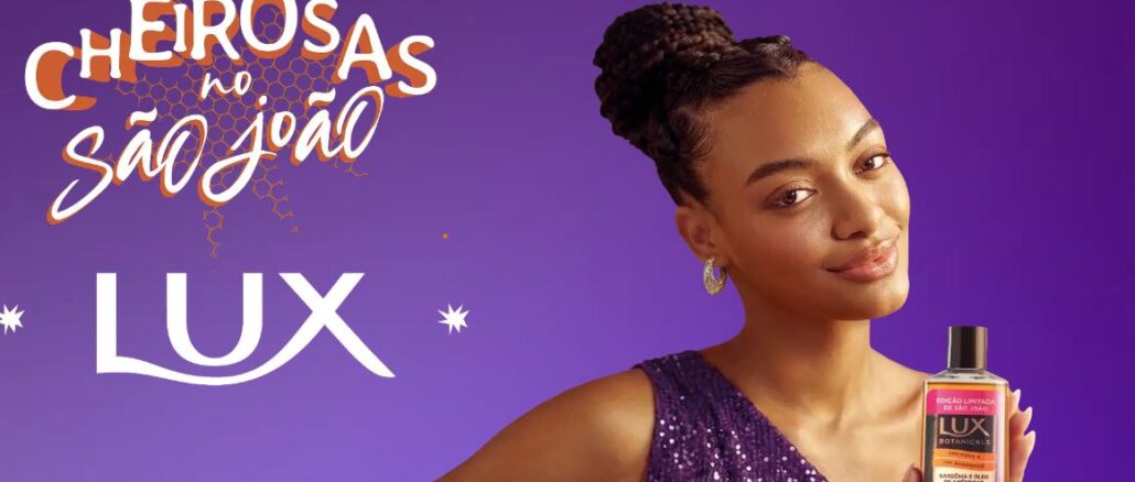 Promoção Cheirosas no São João LUX Imagem promocional da marca Lux com fundo roxo. Do lado esquerdo, lê-se Promoção Cheirosas no São João e o logotipo da marca. Do lado direito, uma mulher com cabelos trançados sorri enquanto segura um frasco de sabonete líquido Lux Botanicals.