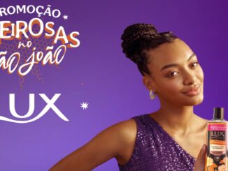 Imagem promocional da marca Lux com fundo roxo. Do lado esquerdo, lê-se Promoção Cheirosas no São João e o logotipo da marca. Do lado direito, uma mulher com cabelos trançados sorri enquanto segura um frasco de sabonete líquido Lux Botanicals.