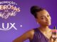 Imagem promocional da marca Lux com fundo roxo. Do lado esquerdo, lê-se Promoção Cheirosas no São João e o logotipo da marca. Do lado direito, uma mulher com cabelos trançados sorri enquanto segura um frasco de sabonete líquido Lux Botanicals.