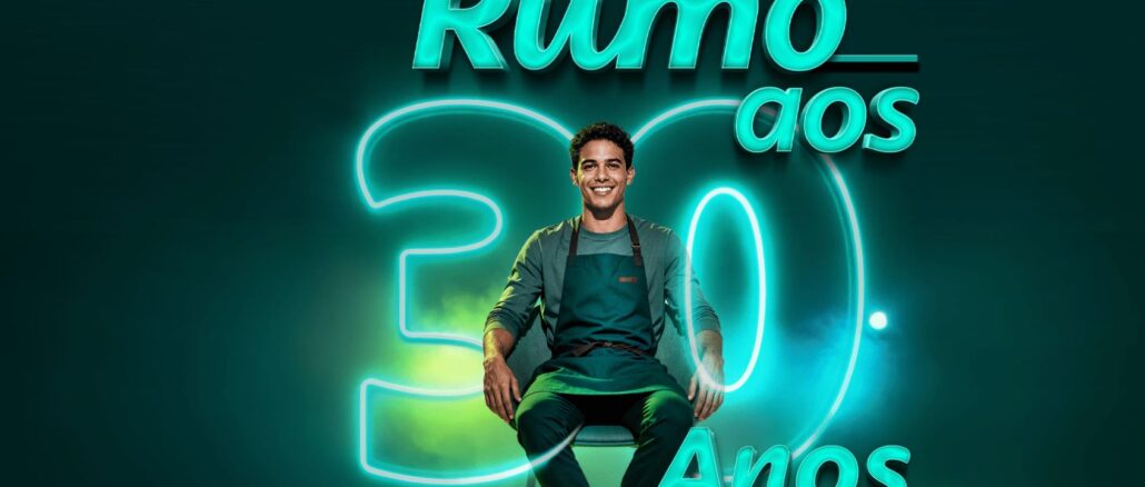 Homem sorridente sentado em frente a um grande número trinta iluminado em neon azul. O banner exibe os textos Promoção Rumo aos 30 Anos Sicoob Divicred em um cenário com tons de verde e fumaça ao fundo.