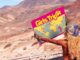 Mulher loira de costas segura um mapa temático do álbum Girls Trip, de Zara Larsson, em uma estrada deserta com montanhas ao fundo.