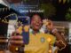 Jovem torcedora com camisa da seleção brasileira segura boneco Quim Torcedor e celular em ambiente decorado com bandeirinhas verde e amarelo, promovendo a Copa Premiada do Quiero Café.