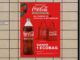Cartaz promocional da Coca-Cola Retornável fixado na parede de um supermercado, anunciando que na compra de 2 unidades do refrigerante retornável o consumidor recebe 1 ecobag vermelha da marca.