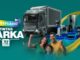 Banner de aniversário de 37 anos da Tintas Darka, em fundo verde-azulado, com caminhão de entrega e diversos eletrodomésticos como geladeira, micro-ondas, lava-e-seca, cafeteira e aspirador robô dispostos em pedestais iluminados, além de uma família formada por pai, mãe e filho sorrindo e comemorando com os punhos erguidos.
