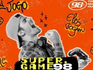 Homem comemora com o punho erguido usando boné e segurando controle de videogame, fundo laranja com ilustrações de bola de futebol e raios, logotipo da 98FM e texto Super Game 98
