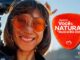 Mulher sorridente usa óculos de sol arredondados com lentes laranja, cabelo castanho ao vento, ao lado de peça em formato de palheta vermelha com o texto Promoção Você e Natura no Rock in Rio 2026, logotipos da Natura e do Rock in Rio no canto superior esquerdo e a frase faz bem estar bem no canto inferior direito