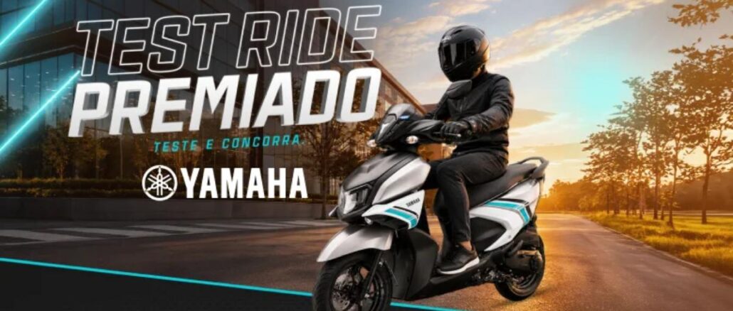 Motociclista com traje preto e capacete integral pilota uma scooter Yamaha branca com detalhes turquesa em uma via arborizada ao entardecer, com prédio espelhado ao fundo e a chamada Test Ride Premiado Yamaha.