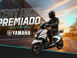 Motociclista com traje preto e capacete integral pilota uma scooter Yamaha branca com detalhes turquesa em uma via arborizada ao entardecer, com prédio espelhado ao fundo e a chamada Test Ride Premiado Yamaha.