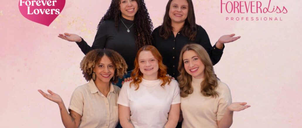 Cinco mulheres sorrindo posam juntas em foto promocional da Comunidade Forever Lovers da Forever Liss Professional, com fundo rosé e confetes dourados.