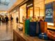 Corredor de shopping center decorado para o Dia dos Pais, com sacolas de presentes coloridas sobre balcão de madeira em primeiro plano e placa "Dia dos Pais – Presentes que Surpreendem" ao fundo.