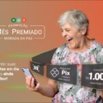 Mulher idosa sorridente segurando celular, ao lado de informações sobre a Promoção Mês Premiado Morada da Paz 2026, que oferece R$1.000,00 por mês a quem pagar contas via Pix.