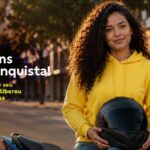 Mulher jovem com cabelos cacheados, usando moletom amarelo e segurando capacete preto, apoiada em moto azul em rua arborizada, com texto "Parabéns pela conquista! A aprovação do seu financiamento liberou uma recompensa" e botão "Saiba Mais".