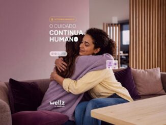 Duas mulheres se abraçam afetuosamente em um sofá, com a frase "O Cuidado Continua Humano" e a marca Wellz by Wellhub sobrepostas à imagem, em campanha do Setembro Amarelo.