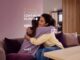 Duas mulheres se abraçam afetuosamente em um sofá, com a frase "O Cuidado Continua Humano" e a marca Wellz by Wellhub sobrepostas à imagem, em campanha do Setembro Amarelo.