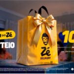 Sacola dourada do Zé Delivery com laço de fita dourada sobre uma mesa, ao lado do texto "Niver do Zé – Sorteio 100 mil em Certificado de Ouro".