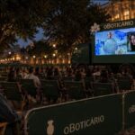 Cinema ao ar livre de O Boticário em praça histórica à noite, com mulher sorridente em cadeira verde, plateia assistindo a tela montada em caminhão e carrinho de pipoca com a inscrição Amor em Cena.
