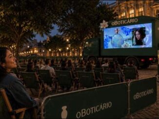 Cinema ao ar livre de O Boticário em praça histórica à noite, com mulher sorridente em cadeira verde, plateia assistindo a tela montada em caminhão e carrinho de pipoca com a inscrição Amor em Cena.