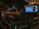 Cinema ao ar livre de O Boticário em praça histórica à noite, com mulher sorridente em cadeira verde, plateia assistindo a tela montada em caminhão e carrinho de pipoca com a inscrição Amor em Cena.