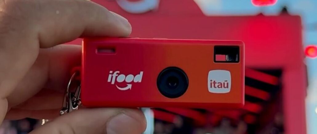 Promoção iFood Itaú RiR Rock In Rio 2026 Câmera fotográfica descartável vermelha com as marcas iFood e Itaú, sendo segurada por uma mão em frente a uma estrutura decorativa vermelha, no contexto do Rock in Rio 2026.