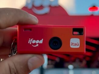 Câmera fotográfica descartável vermelha com as marcas iFood e Itaú, sendo segurada por uma mão em frente a uma estrutura decorativa vermelha, no contexto do Rock in Rio 2026.
