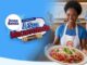 Mulher sorridente com avental do MasterChef Brasil segura um prato de macarronada com molho de tomate, queijo ralado e manjericão, ao lado do logotipo da Promoção A Sua Macarronada Dona Benta, com um homem e uma menina comemorando ao fundo em uma cozinha moderna.