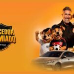 Arte da promoção Torcedor Premiado Vale Bônus com Marcos Mion sorridente apontando para um celular, ao lado de três mascotes cachorros vestindo camisas de futebol, um carro sedã dourado e uma pilha de barras de ouro sobre fundo laranja.