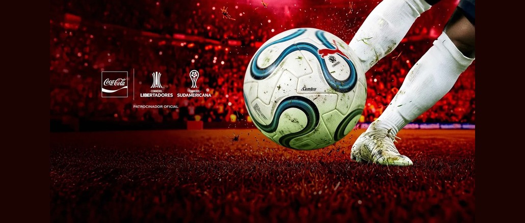 Promoção Coca-Cola Bola do Jogo Bola de futebol branca e azul com a marca Cumbre sendo chutada por um jogador de meiões brancos, em um estádio lotado iluminado em vermelho, ao lado dos logotipos da Coca-Cola, da Conmebol Libertadores e da Conmebol Sudamericana.