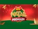 Logotipo Natal Premiado em letras douradas sobre coroa natalina com laço vermelho e sinos dourados, faixa vermelha com a palavra Premiado, fundo vermelho com estrelas douradas e chão verde