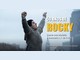 Cena de "Eu Sou o Rocky" com o ator Anthony Ippolito, como Sylvester Stallone, de braços erguidos e mãos enfaixadas, com o texto "50 anos de Rocky: conte sua história e concorra a 1 de 5 kits".