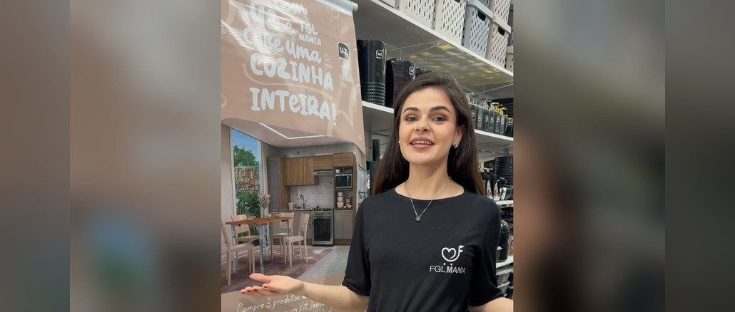 Com UZ e FGL Cabe uma Cozinha Inteira! Mulher com camiseta preta da FGL Mania sorri ao lado de banner da promoção "Com UZ e FGL Mania cabe uma cozinha inteira!", com foto de cozinha planejada e mesa com cadeiras, em loja com prateleiras de utilidades UZ.