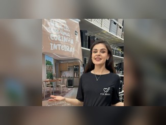 Mulher com camiseta preta da FGL Mania sorri ao lado de banner da promoção "Com UZ e FGL Mania cabe uma cozinha inteira!", com foto de cozinha planejada e mesa com cadeiras, em loja com prateleiras de utilidades UZ.