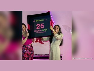 Duas mulheres seguram uma grande caixa preta com laços rosa que exibe a mensagem "Ele voltou 25 Prêmio Avon de Maquiagem", em um cenário rosa com luzes de neon.